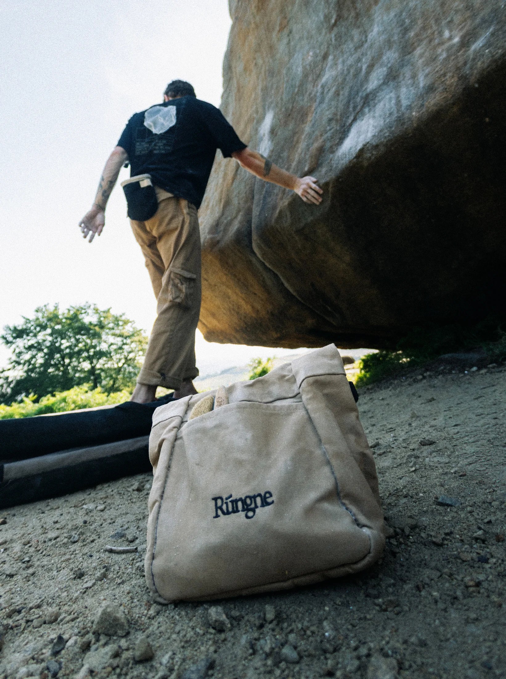 A Rúngne canvas chalk bucket open on gritstone in the foreground; above it a climber reaches across a boulder.