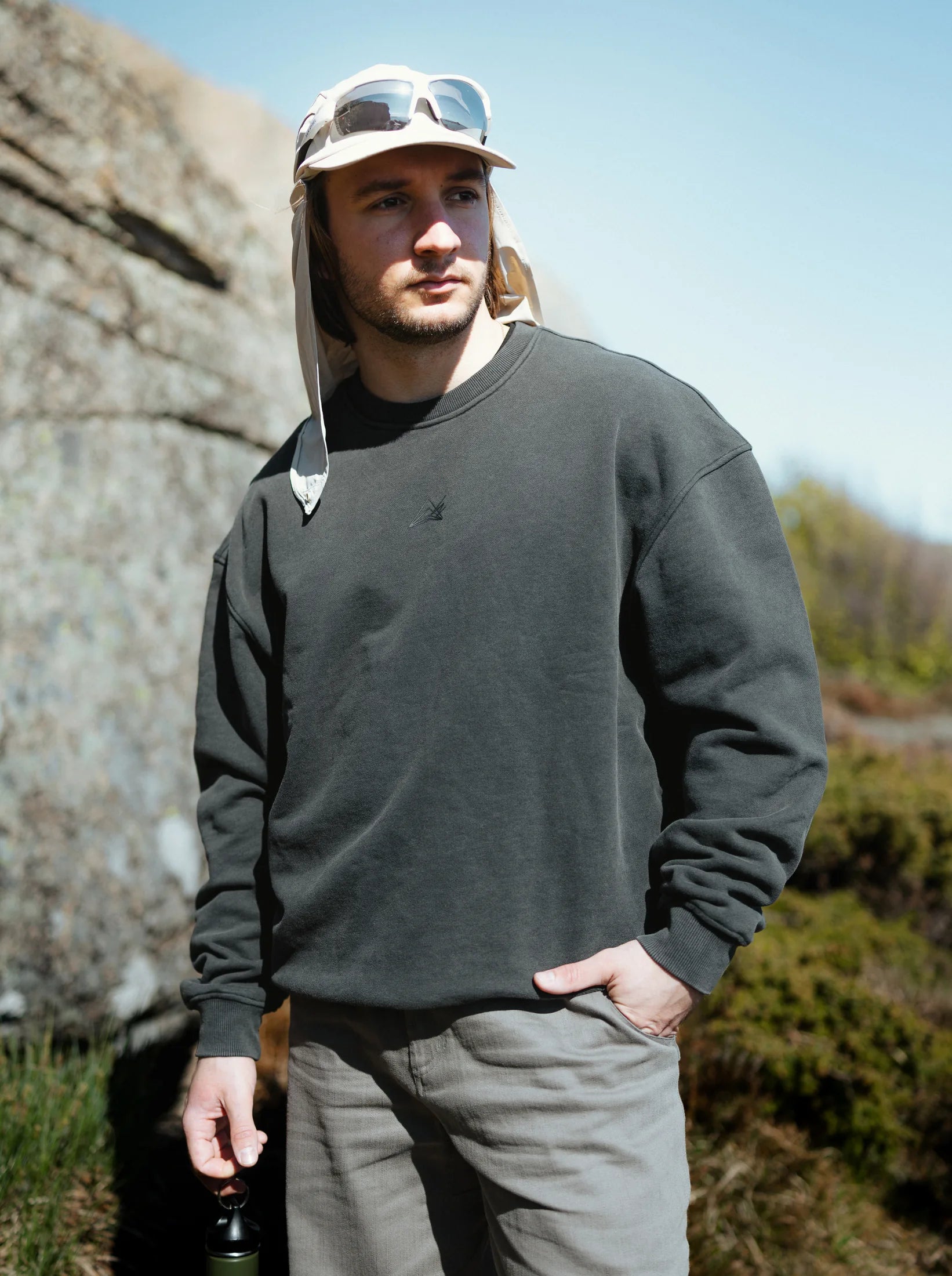 A climber stands on coastal heath in a charcoal Project Crewneck and grey trousers, a granite boulder behind.