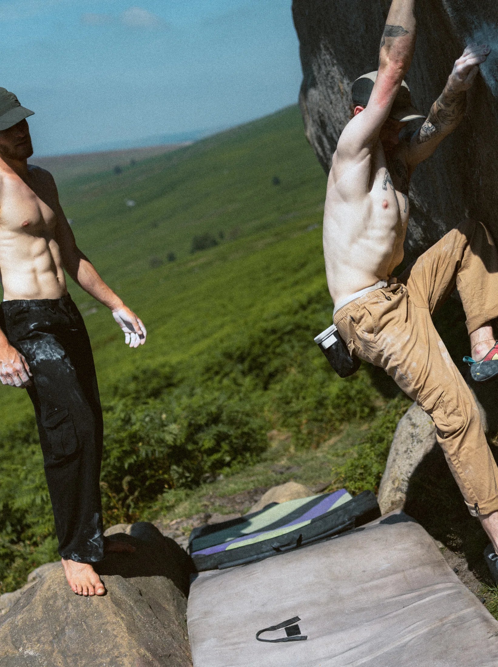 A climber stands on a bouldering pad on open moorland, another working the boulder behind.