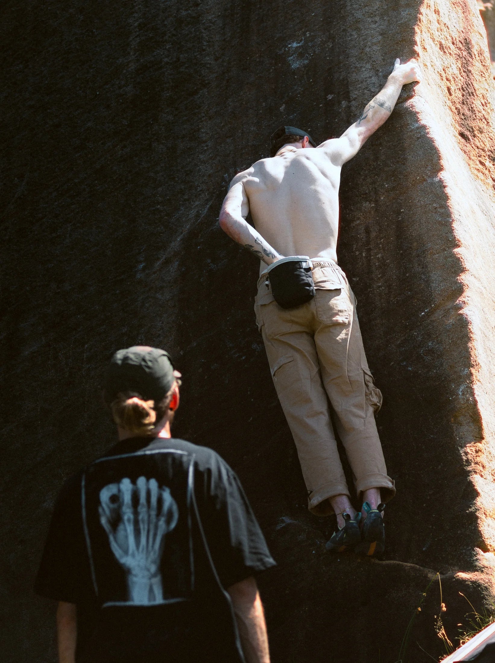 Two climbers on a gritstone buttress, one watching from below in a printed tee.