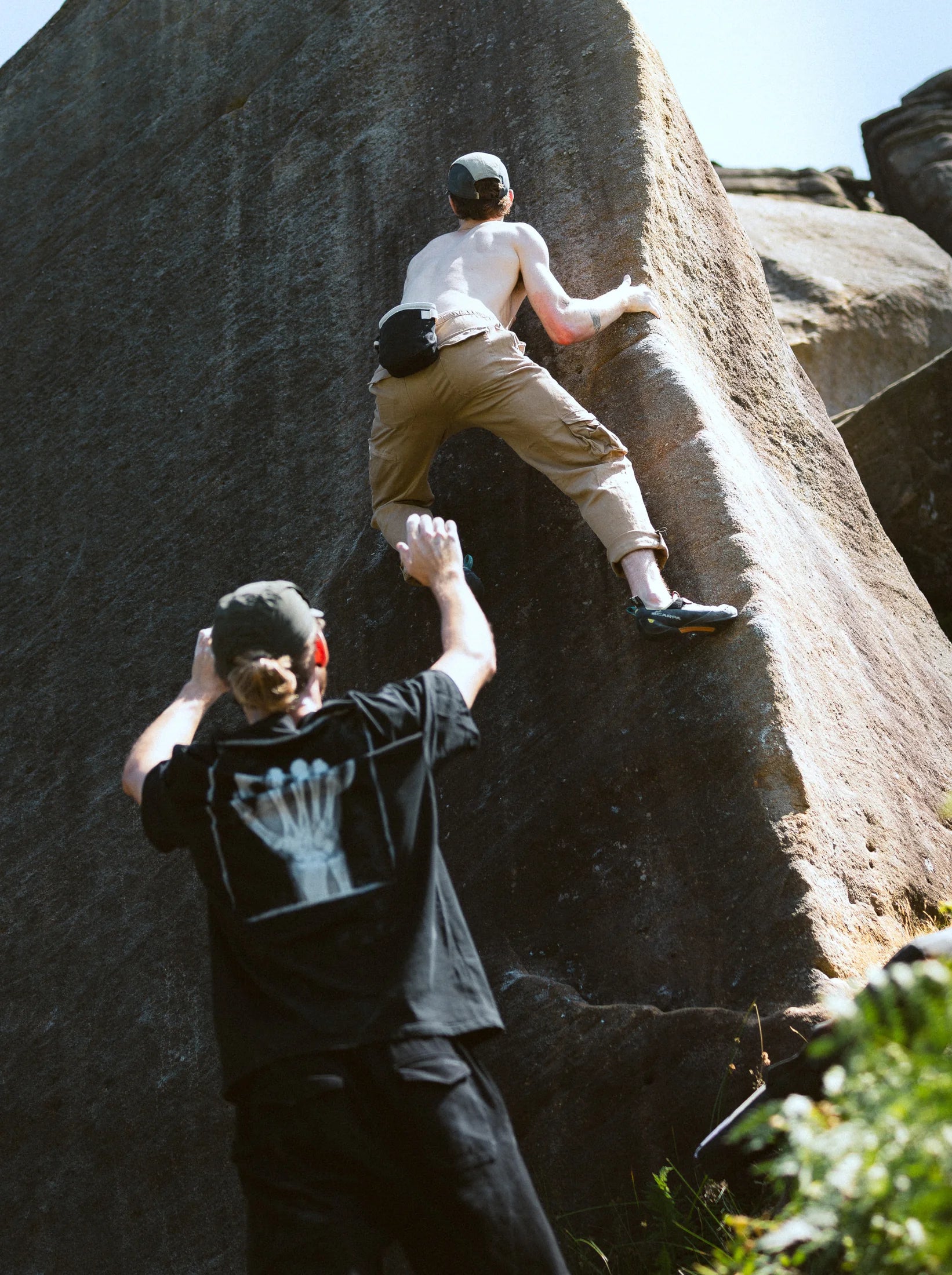 A climber pulls over a gritstone bulge in a printed tee, a second figure watching from the shade.